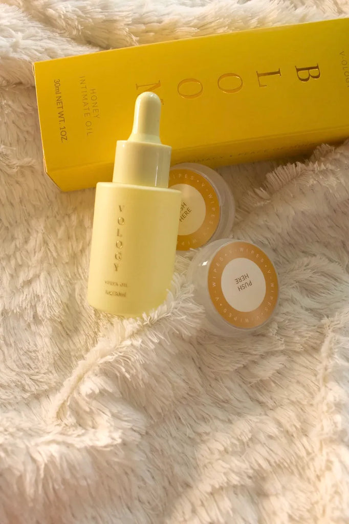 A Bloom Honey Intimate Oil bottle with a yellow box on a white blanket, designed to address vaginal odor.
